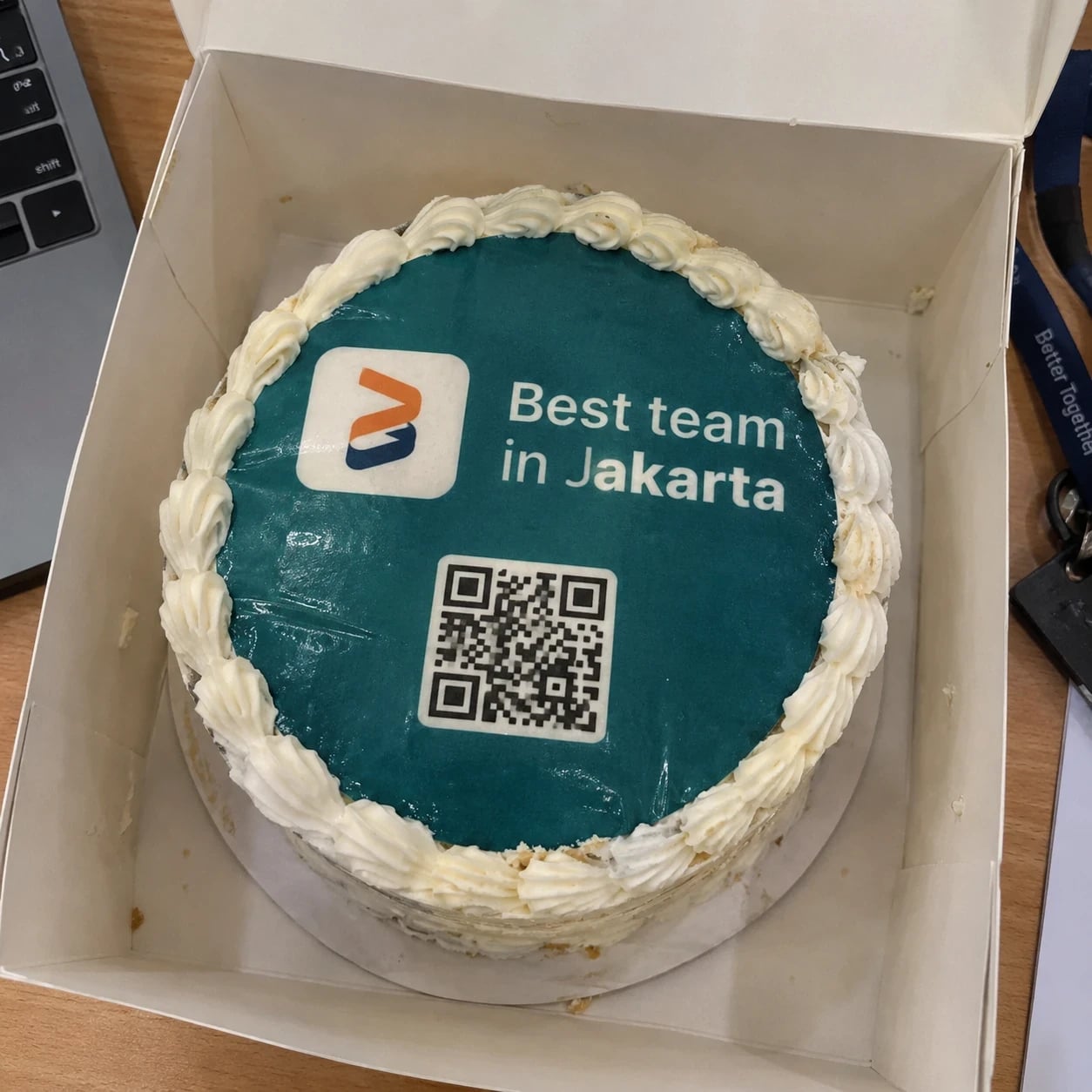 An open cake box with a cake reading “Best team in Jakarta”, a logo and a QR code