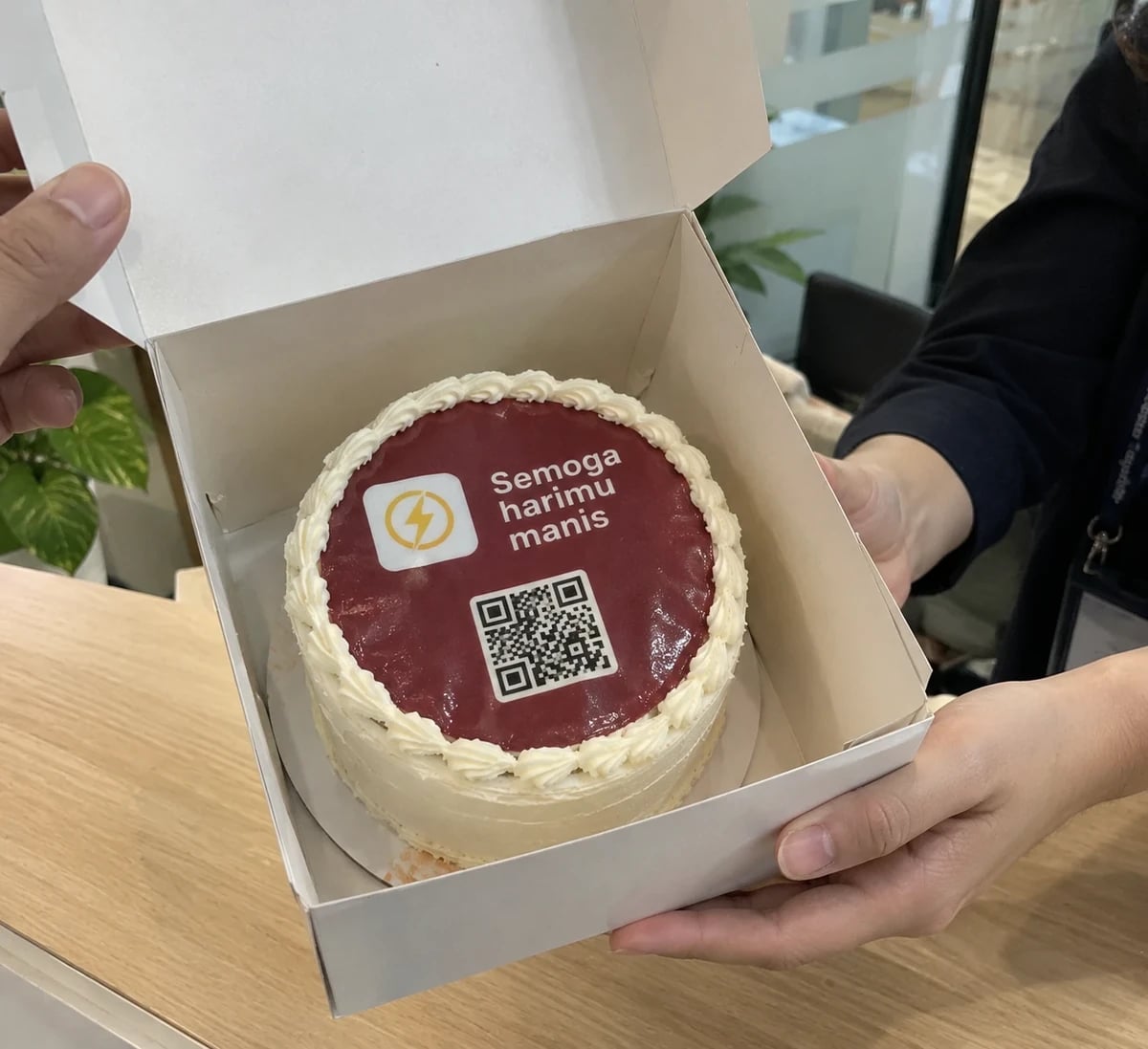 An open cake box in hand, the cake reading “Semoga harimu manis”